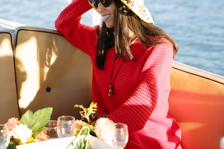 Woman in a red sweater and hat smiling on a boat with fruit platter nearby.