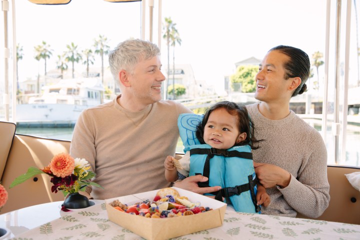 Two men and a child in a blue life jacket smiling on a boat with flowers and food box on table.