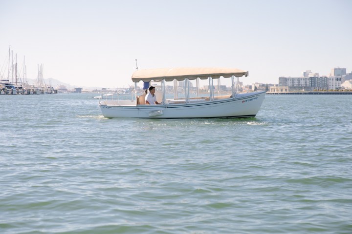 Small covered boat on water with a person onboard, city and marina in the background.