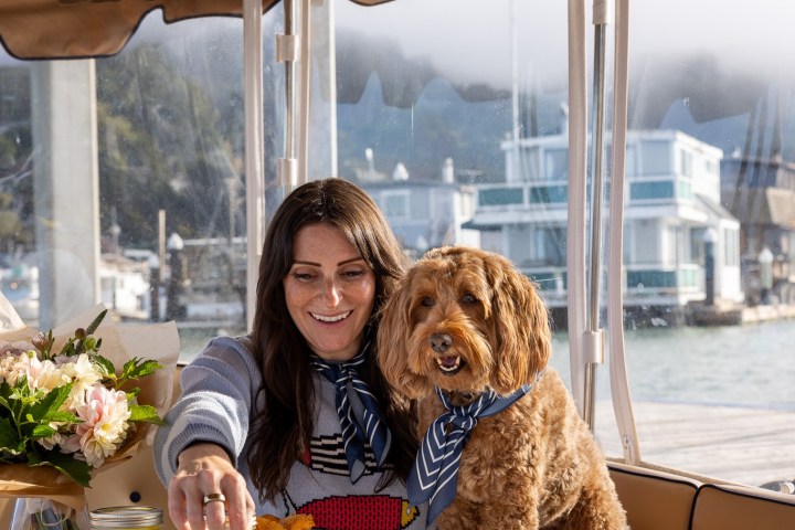 Woman and dog on a boat, enjoying a meal with salads and fries, surrounded by flowers.