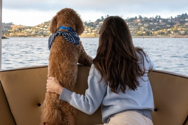 Woman and dog with scarf look out from boat at lake and hills.