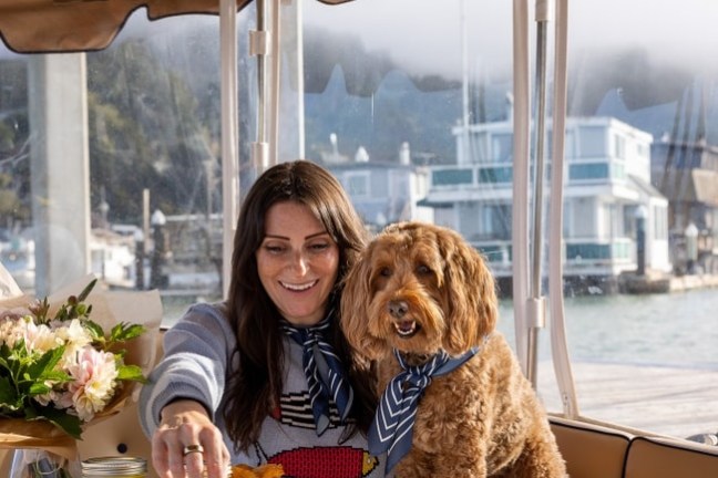 Woman with dog enjoying food on a boat, table surrounded by water and buildings.