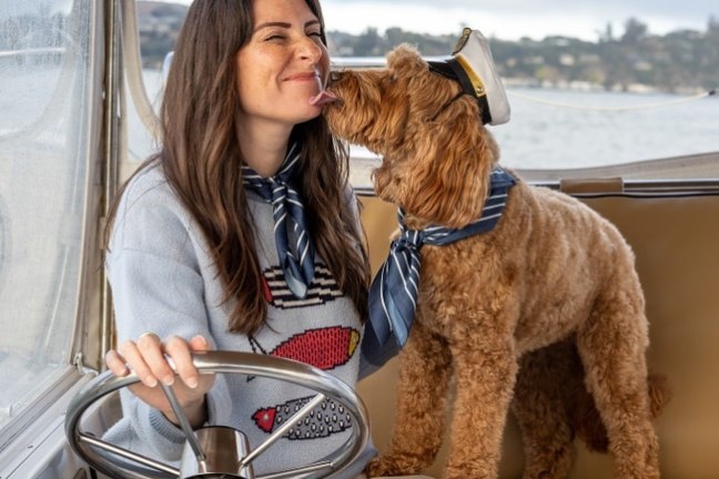 Woman smiling at dog wearing a sailor hat on a boat.