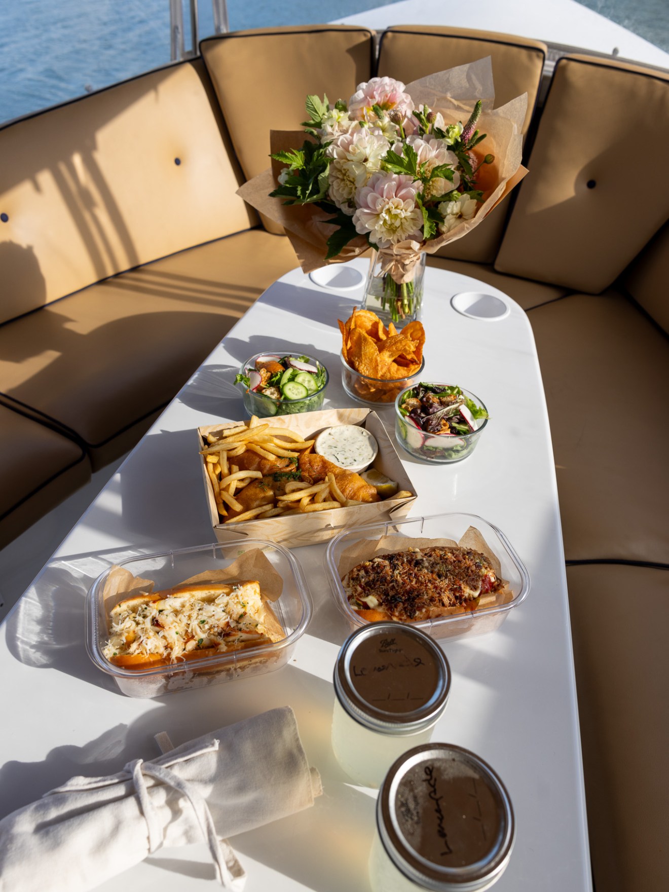 Table with flowers, sandwiches, fries, salads, and drinks on a boat.