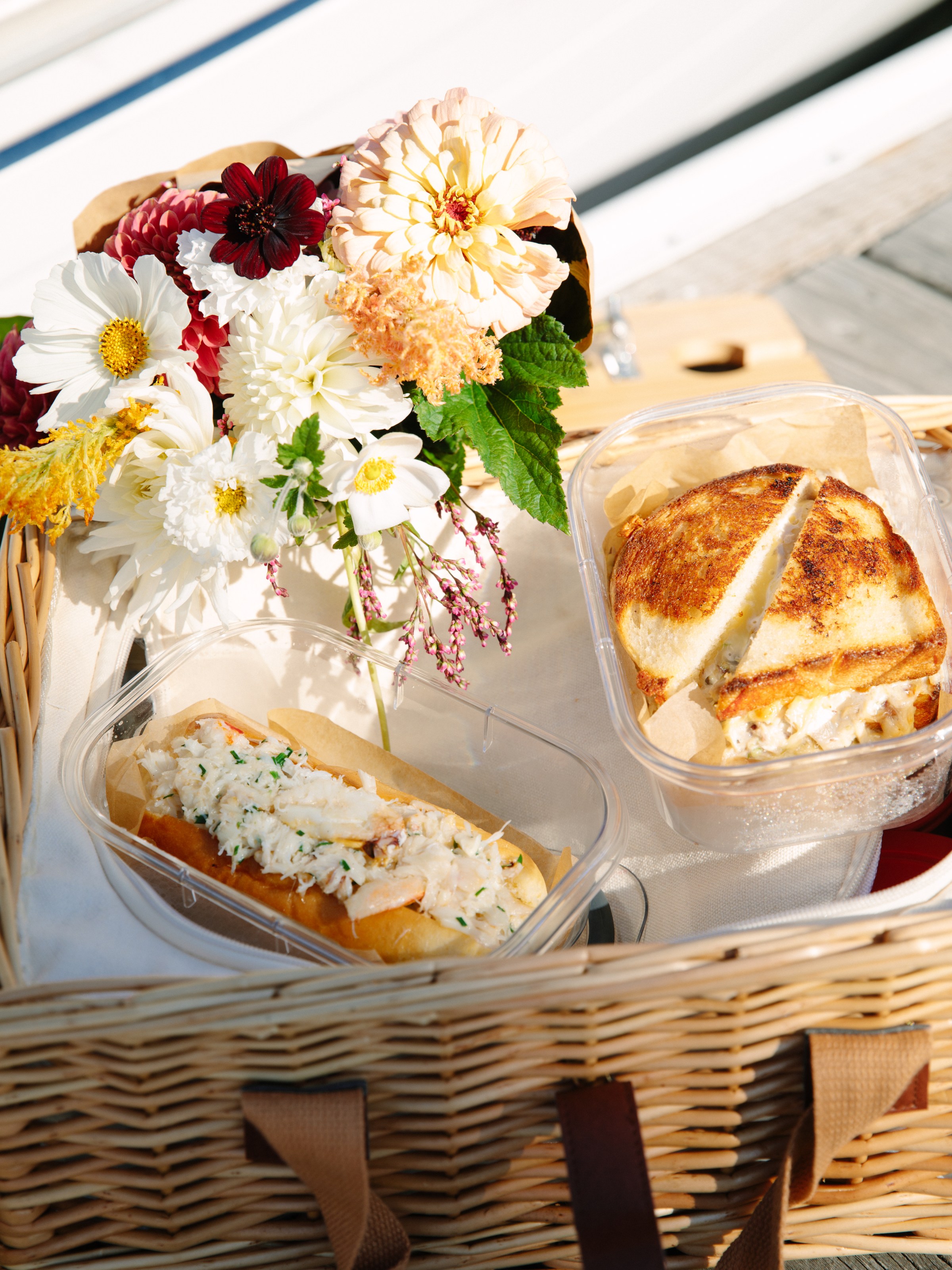 Picnic basket with sandwiches and a bouquet of colorful flowers on a wooden surface.