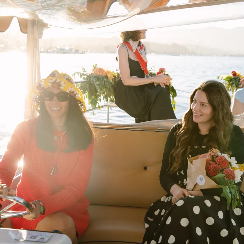 Women on a boat; one steers while another holds flowers, others sit in background with floral decorations.