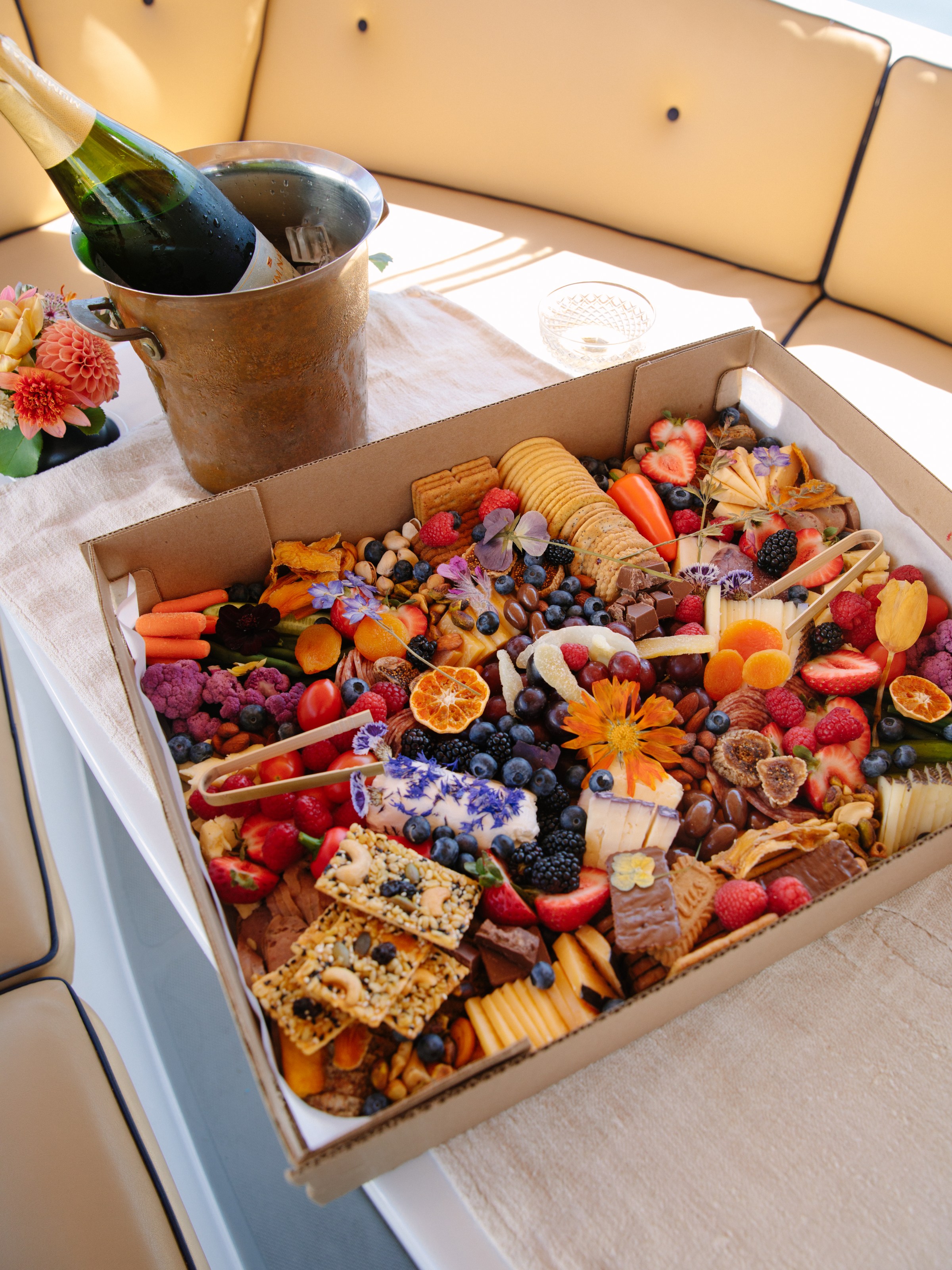 Charcuterie board with fruits, cheeses, and crackers next to a bucket with a bottle on a boat's seating area.