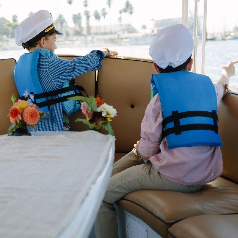 Two children in captain hats and life vests sit on a boat, with flowers on the table.